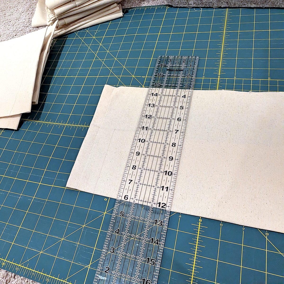 A clear quilting ruler laid across a cut panel of natural canvas on a green cutting mat, with a stack of cut panels behind it.