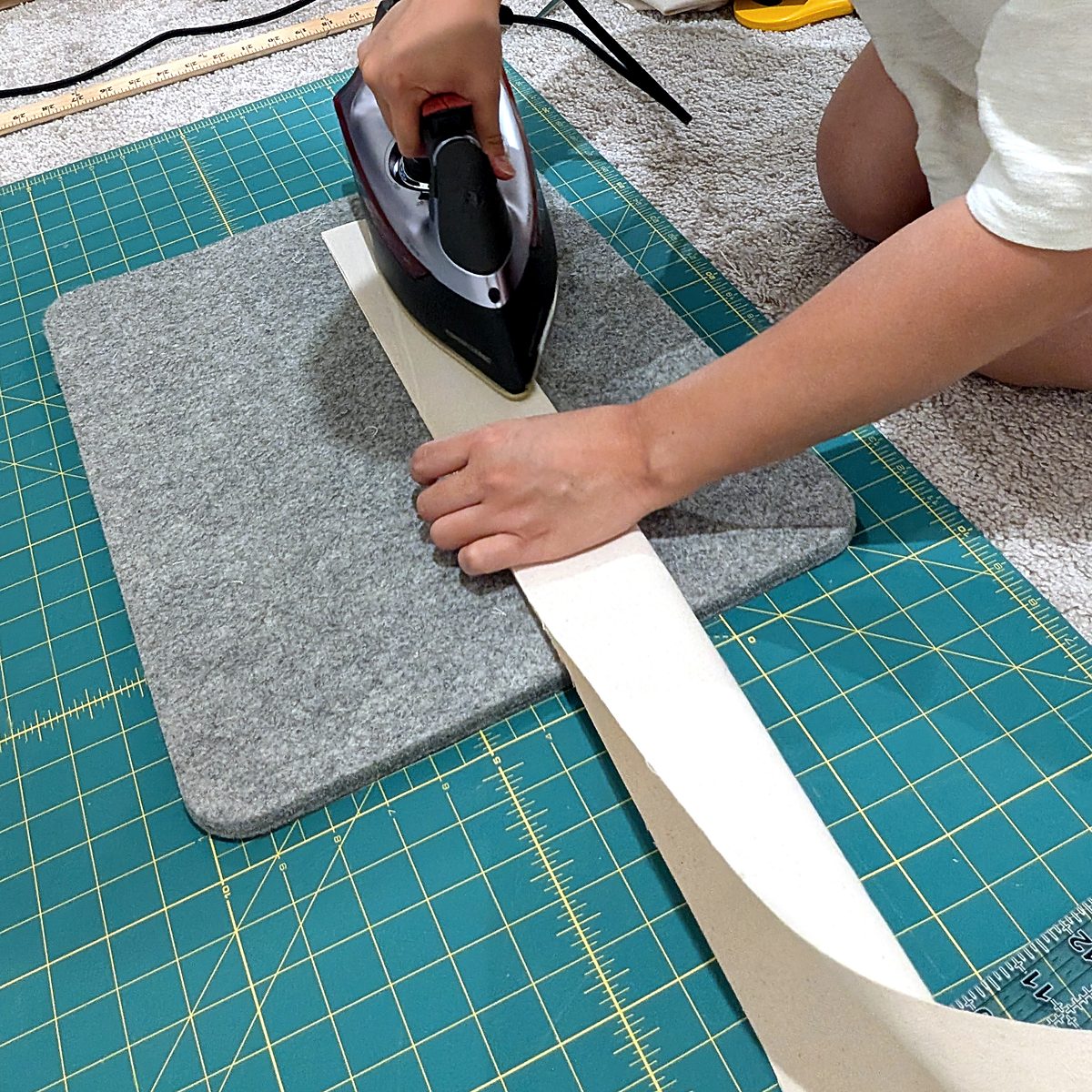 Hands pressing a long canvas strap flat with an iron on a gray wool pressing mat.