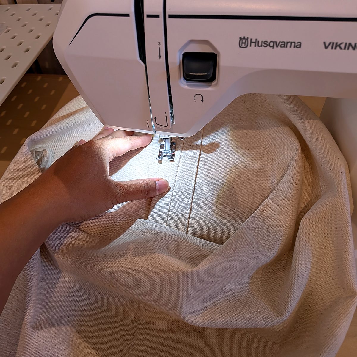 Hands guiding the canvas bag body under the presser foot of a white home sewing machine, topstitching a seam.