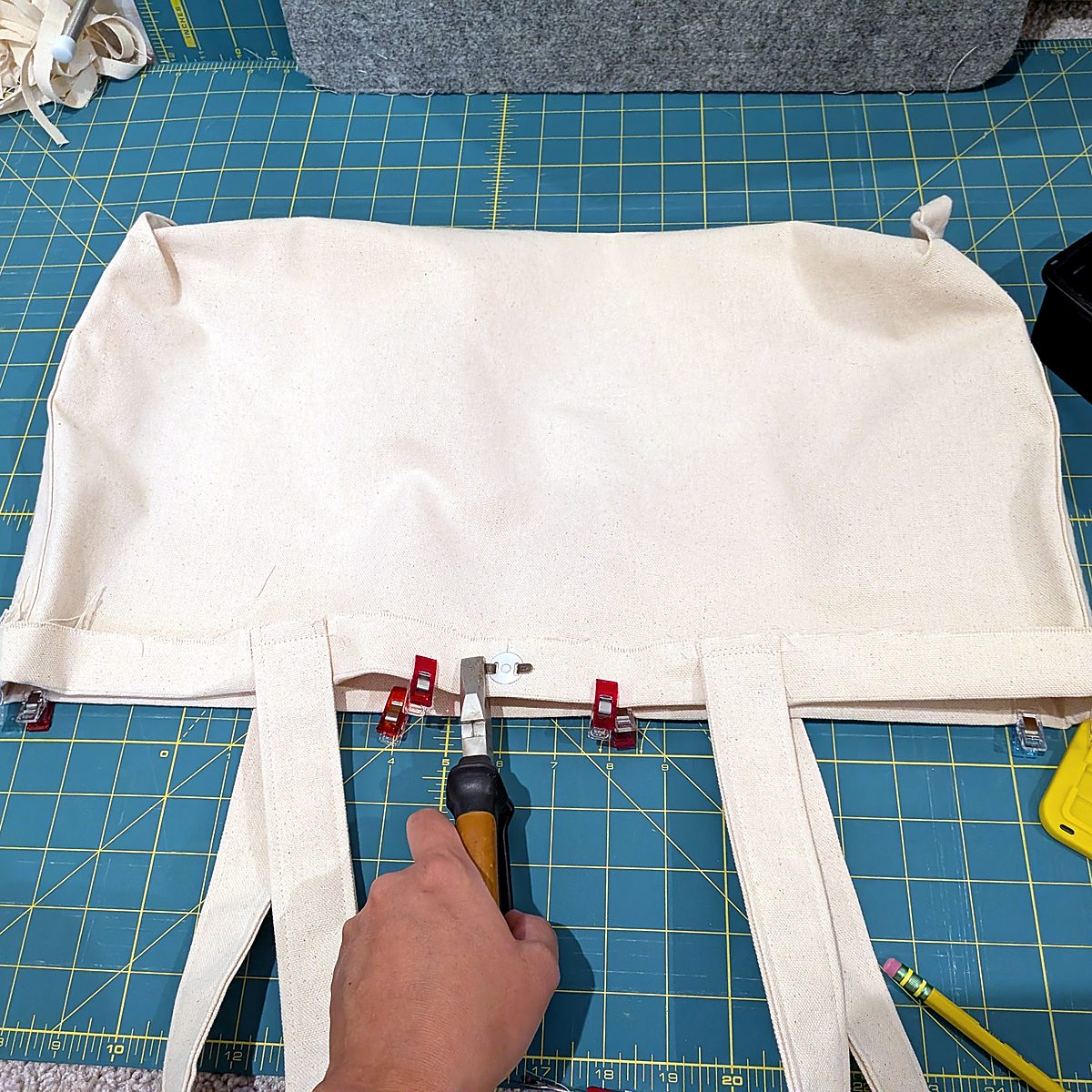 Overhead view of a nearly finished tote on a cutting mat, clipped along the top edge, as a magnetic snap is set by hand with pliers.