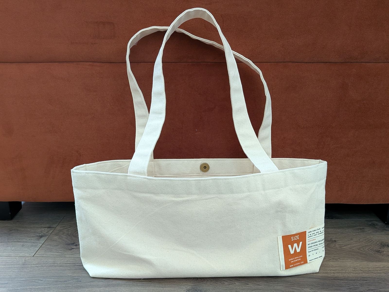The SIZE WIDE tote standing on a wood floor, wide and shallow, with its handles resting back over the rim.
